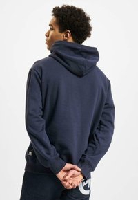 Navy blue hoodie with a drawstring hood, ribbed cuffs, and hem. Soft texture, featuring a logo patch near the hem. Simple, classic design.