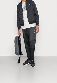 Zwarte bomberjack, grijs t-shirt, zwarte broek met zakken, zwarte sneakers en een grijze rugzak. Heeft Nike-logo's op kleding en rugzak.