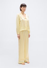 Γυναίκα που στέκεται ξυπόλυτη φορώντας ανοιχτό κίτρινο σατέν σετ πιτζάμες με μεγάλο γιακά και κουμπιά, παντελόνια με φαρδιά μπατζάκια και μπλε περιθώριο στα μανίκια.