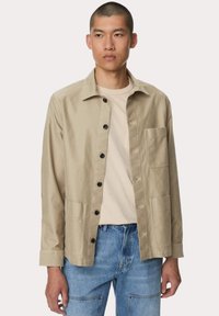 Beige katoenen overhemd met knopen en een kraag, twee voorzakken en zwarte knopen. Draag het over een lichtbeige t-shirt en blauwe spijkerbroek.
