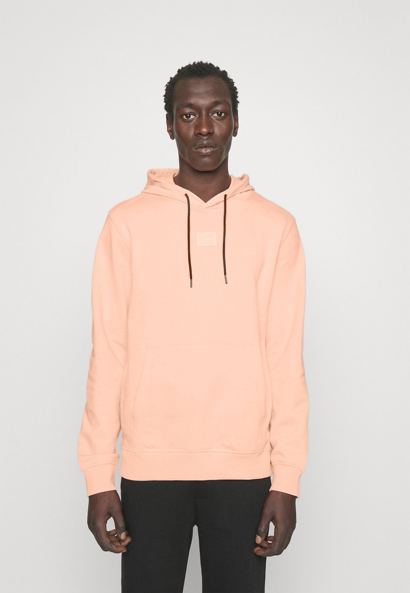 Mikina s kapucí ve měkké broskvové látce s přední kapsou, šňůrkou a čtvercovou nášivkou loga, v kombinaci s černými kalhotami.