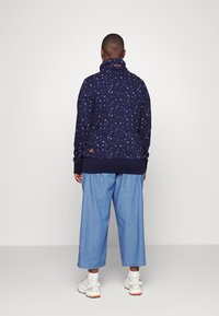 Suéter de patrón marino con cuello alto, que presenta pequeños diseños multicolores. Combinado con pantalones de pierna ancha de color azul claro y zapatillas blancas.