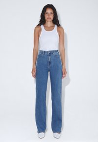 Jeans in denim blu a vita alta con taglio dritto, dotati di cinque tasche e chiusura con bottone. Indossati con un top bianco senza maniche.