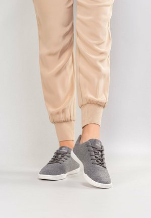 Persoon die beige joggerbroek en grijze sportschoenen met witte zolen draagt, staat op een lichtgrijze ondergrond.