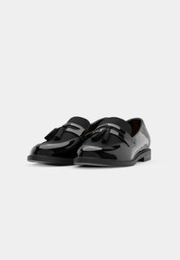 Scarpe in vernice nera con suola piatta, caratterizzate da un accento decorativo a nappina e un design semplice. Texture liscia e forma arrotondata.