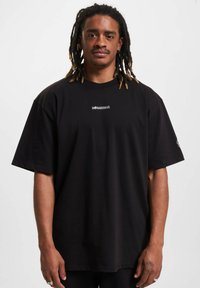 Juodas per didelis medvilninis marškinėliai su apvalia iškirpte ir trumpomis rankovėmis. Yra mažas logotipas atspausdintas ant krūtinės ir paprastas dizainas.