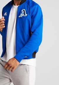 Cazadora bomber atlética azul con cierre frontal, puños acanalados y un logo bordado en el pecho. Presenta bolsillos laterales y una textura suave.