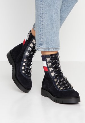 Bottines en daim bleu marine à lacets avec semelles épaisses noires, ferrures argentées, détail rayé rouge et blanc, portées avec un jean bleu clair.