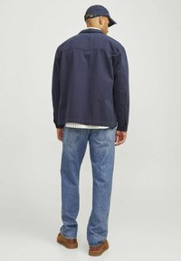 Chaqueta de algodón azul marino con un corte relajado, costuras visibles, combinada con jeans de mezclilla azul claro y zapatos marrones. Vista trasera.