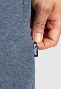 Pantalones de tela gris-azul, con una pequeña etiqueta negra con texto blanco, mostrando parte de una mano sujetando la etiqueta. Material suave y texturizado.