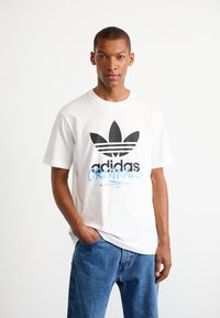 T-shirt di cotone bianco con grande logo adidas nero e testo blu "ORIGINALS". Abbinato a jeans in denim azzurro chiaro.