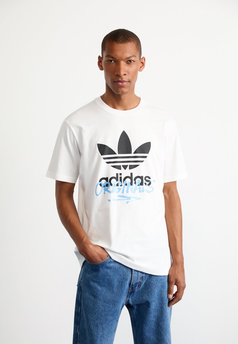 T-shirt di cotone bianco con grande logo adidas nero e testo blu "ORIGINALS". Abbinato a jeans in denim azzurro chiaro.