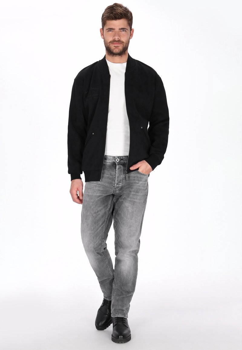 Mann trägt schwarze Bomberjacke, weißes Hemd, grau verwaschene Jeans und schwarze Stiefel, steht mit einer Hand in der Tasche vor weißem Hintergrund.