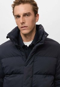 Chaqueta de plumón azul marino con cuello alto, presenta un cierre de cremallera y textura acolchada. Llevada sobre una camiseta de polo oscura.