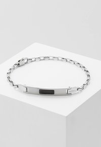Pulsera de cadena de metal plateado con un panel de acento negro texturizado y un cierre seguro. El diseño es elegante y moderno, con un logotipo de la marca integrado.