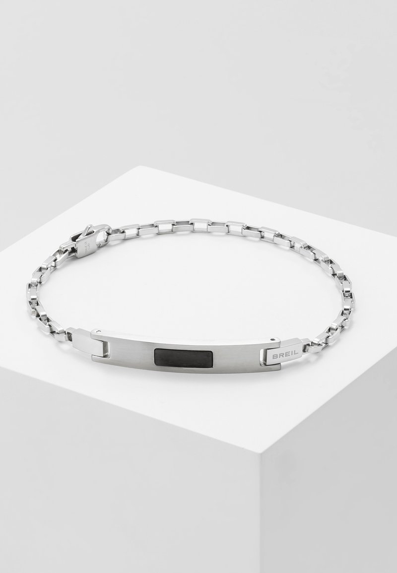 Pulsera de cadena de metal plateado con un panel de acento negro texturizado y un cierre seguro. El diseño es elegante y moderno, con un logotipo de la marca integrado.