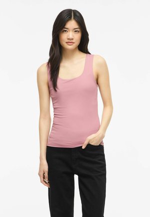 Jeune femme aux longs cheveux foncés portant un haut rose sans manches et un pantalon noir sur un fond blanc uni.