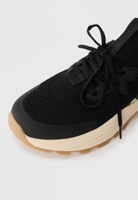 Sneaker atletico nero con tomaia in mesh, suola in gomma e lacci neri piatti. La suola presenta un accento beige con motivi testurizzati.