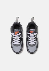 Nike tornacipők szürke és fekete dizájnnal, fehér fűzőkkel, húzófüllel és egy kiemelkedő narancssárga logóval a nyelvén. Vastag talp.