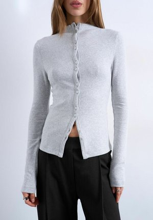 Kvinde iført en lysegrå, langærmet skjorte med nogle knapper åbne, kombineret med sorte bukser.