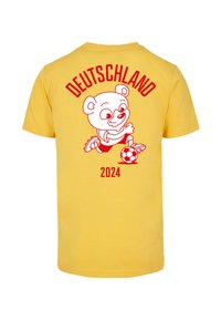 Žluté bavlněné tričko s obrázkem červeného kresleného medvěda, který hraje fotbal. Nápis "DEUTSCHLAND 2024" je nad obrázkem medvěda.