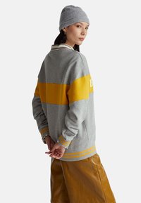 Donna con berretto grigio, maglione a righe grigie e gialle e gonna in pelle marrone, di profilo con le mani intrecciate dietro la schiena.