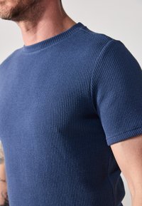 Blå ribbestrikket T-skjorte med rund hals og korte ermer. Teksturen er jevn med vertikale linjer, som viser en tettsittende design.