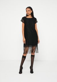 Melna mežģīņu kleita ar īsām piedurknēm, bārkstīm apakšmalā un brīvu piegriezumu. Modele valkā caurspīdīgas melnas augstās zeķubikses līdz augšstilbiem un augstpapēžu kurpes.