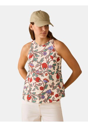 Donna che indossa una canotta floreale senza maniche rossa, viola e verde su sfondo crema, pantaloni color crema e un berretto da baseball beige, che guarda di lato.