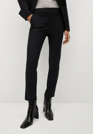 Femme portant un pantalon noir slim, des bottines noires à talons et un blazer noir avec les mains dans les poches.