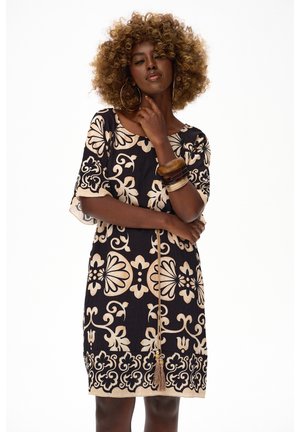 Kobieta o kręconych włosach, ubrana w czarną sukienkę z beżowymi kwiatowymi wzorami, duże kolczyki-koła i kilka bransoletek, pozująca z ręką na brodzie.