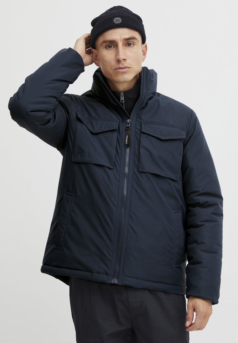 Dunkelblaue isolierte Jacke mit Reißverschluss, hohem Kragen und zwei Brusttaschen. Glatte Textur und lockere Passform. Schwarze Strickmütze.