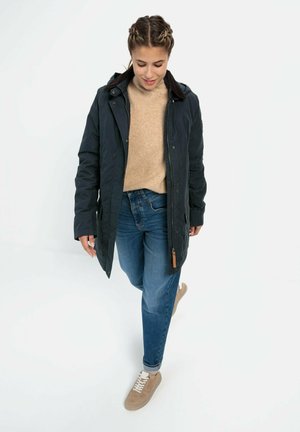 Dunkelblau gesteppter Mantel mit Kapuze, braunem Kragen, beigem Pullover und blauen Jeans. Beigefarbene Sneaker mit weißen Schnürsenkeln, leichtem Material und lässiger Passform.
