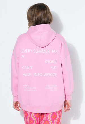 Ruskeatukkainen henkilö pukeutuneena vaaleanpunaiseen huppariin, jossa on valkoista tekstiä, ja värikkäisiin kuvioituihin housuihin, seisoo valkoisen seinän vieressä selin päin.