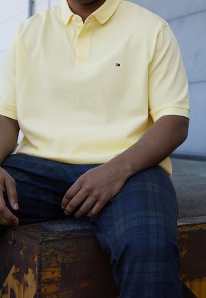 Gelbes Polohemd aus Baumwolle mit einer Knopfleiste mit zwei Knöpfen und einem kleinen Logo. Kombiniert mit dunkel karierten Hosen mit blauen Akzenten.