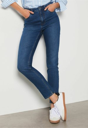 Donna che indossa jeans skinny blu scuro a vita alta e sneakers bianche, mani nelle tasche, in piedi con le gambe incrociate appoggiata al muro.