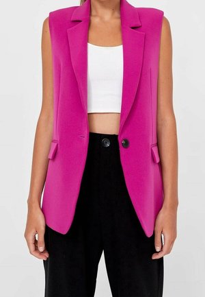 Woman wearing a bright pink sleeveless blazer over a white cropped top and black high-waisted pants with buttons.