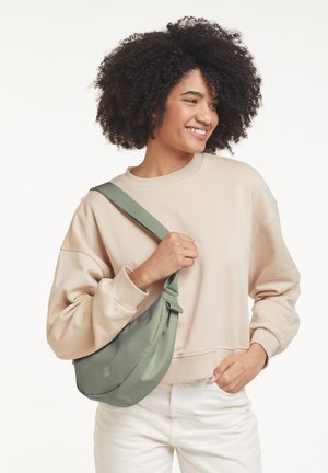 Groene nylon schoudertas met een knoopdetail, gladde textuur en logo. Ggedragen met een beige sweatshirt en witte broek.