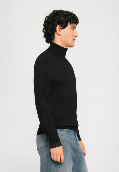 Mann im schwarzen Rollkragenpullover und blauer Jeans steht mit dem Gesicht nach rechts vor einem schlichten hellen Hintergrund.