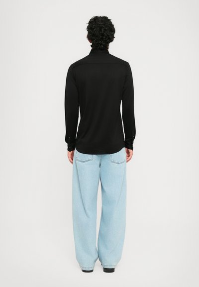 En person med krøllet hår står med ryggen til, iført en svart langermet skjorte og lyseblå vide jeans mot en ensfarget bakgrunn.