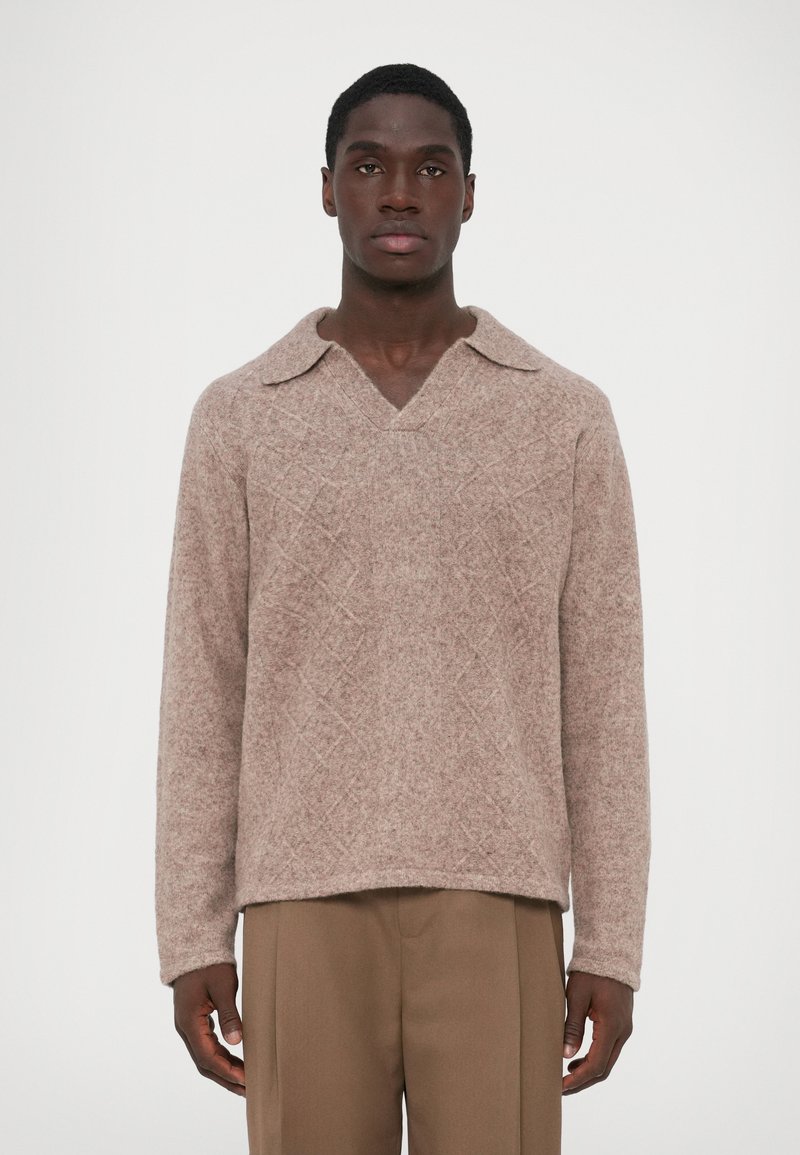 Pull en tricot marron clair avec un col large, motif en diamant texturé, et manches longues, associé à un pantalon beige.