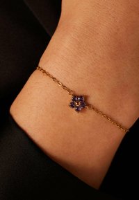 Gouden kettingarmband met een bloemontwerp en paarse edelstenen in het midden. De textuur is glad en de algehele vorm is delicaat.