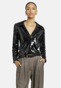 Schwarze Paillettenbluse mit Kragen und langen Ärmeln, die eine glänzende Textur aufweist, kombiniert mit hoch taillierten, gefalteten beigen Hosen.