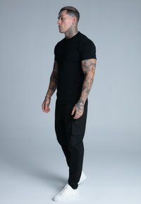 Camiseta negra ajustada, pantalones cargo negros con bolsillos laterales y zapatillas blancas. El modelo tiene tatuajes visibles en los brazos, posando de perfil.