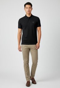 Svart pikétröja med ett subtilt geometriskt mönster, korta ärmar och en klassisk krage, kombinerad med ljusbeige byxor och bruna loafers.