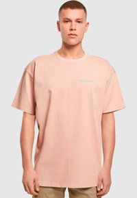 Světle růžové oversized tričko vyrobené z bavlny, s kulatým výstřihem a krátkými rukávy, s jemným modrým textem na přední straně.