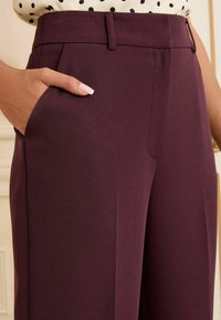 Pantalons bordeaux en tissu texturé, avec des poches latérales, une taille plate et une coupe ajustée. Lignes épurées et coutures simples.