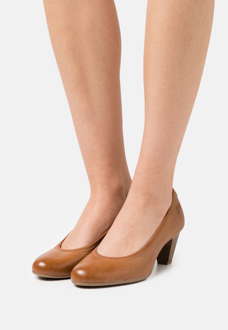 Jambes portant des chaussures en cuir marron à talons bas fermés à l’avant, avec des talons empilés, sur un fond clair uni.