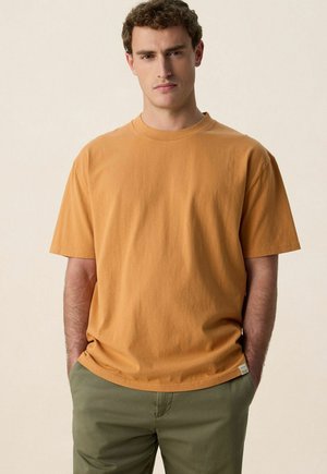 Homme aux cheveux bouclés portant un t-shirt à manches courtes couleur moutarde et un pantalon vert olive, debout les mains dans les poches.