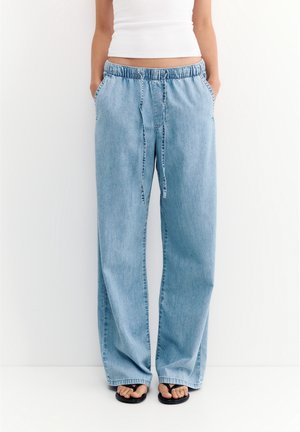 Personne portant un pantalon en jean bleu clair ample avec une taille élastique et un cordon, un débardeur blanc court, et des sandales noires flip-flop.